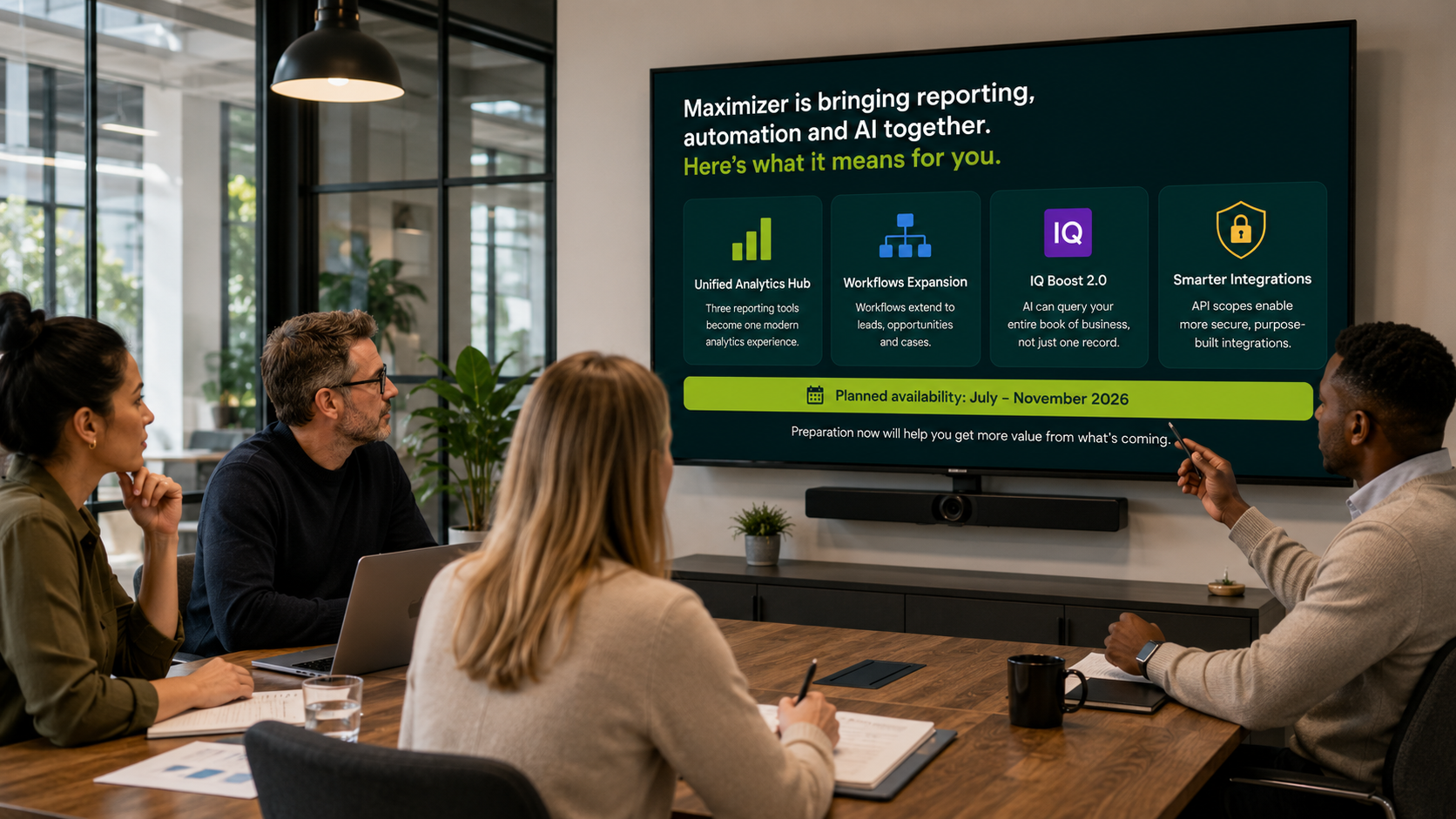 Image shows four people sat around a meeting table, discussing Maximizer's latest updates on the TV screen